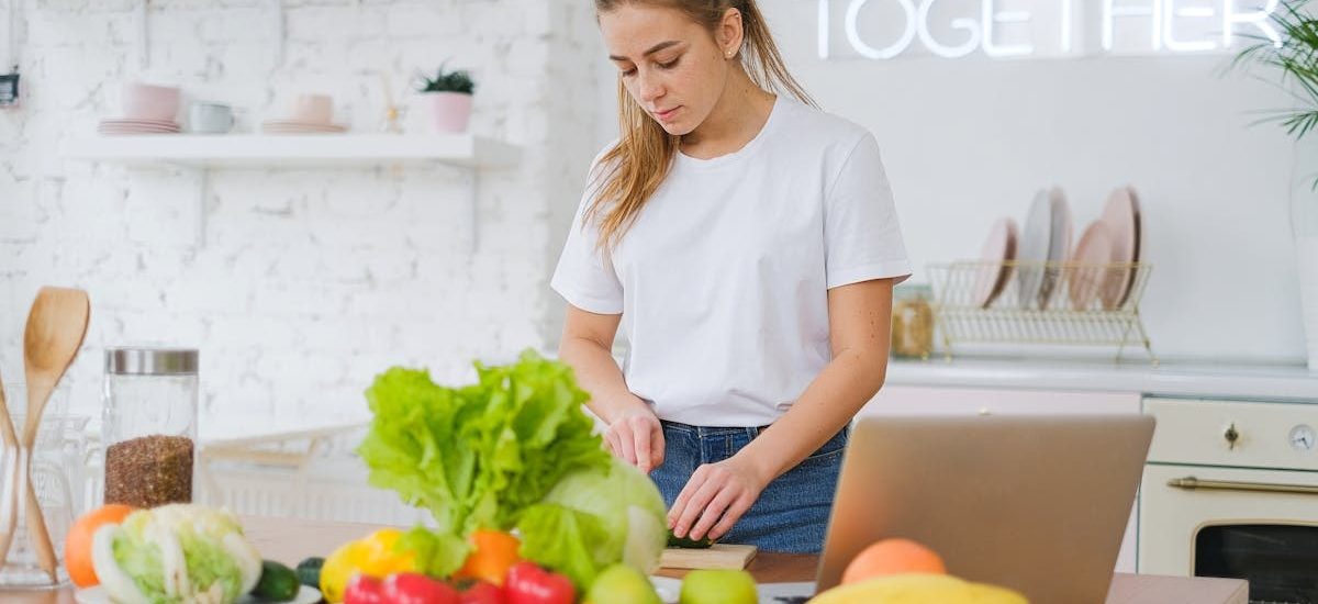 Woman in white t-shirt preparing fresh vegetables in a bright kitchen with a laptop.
