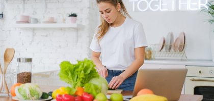 Woman in white t-shirt preparing fresh vegetables in a bright kitchen with a laptop.